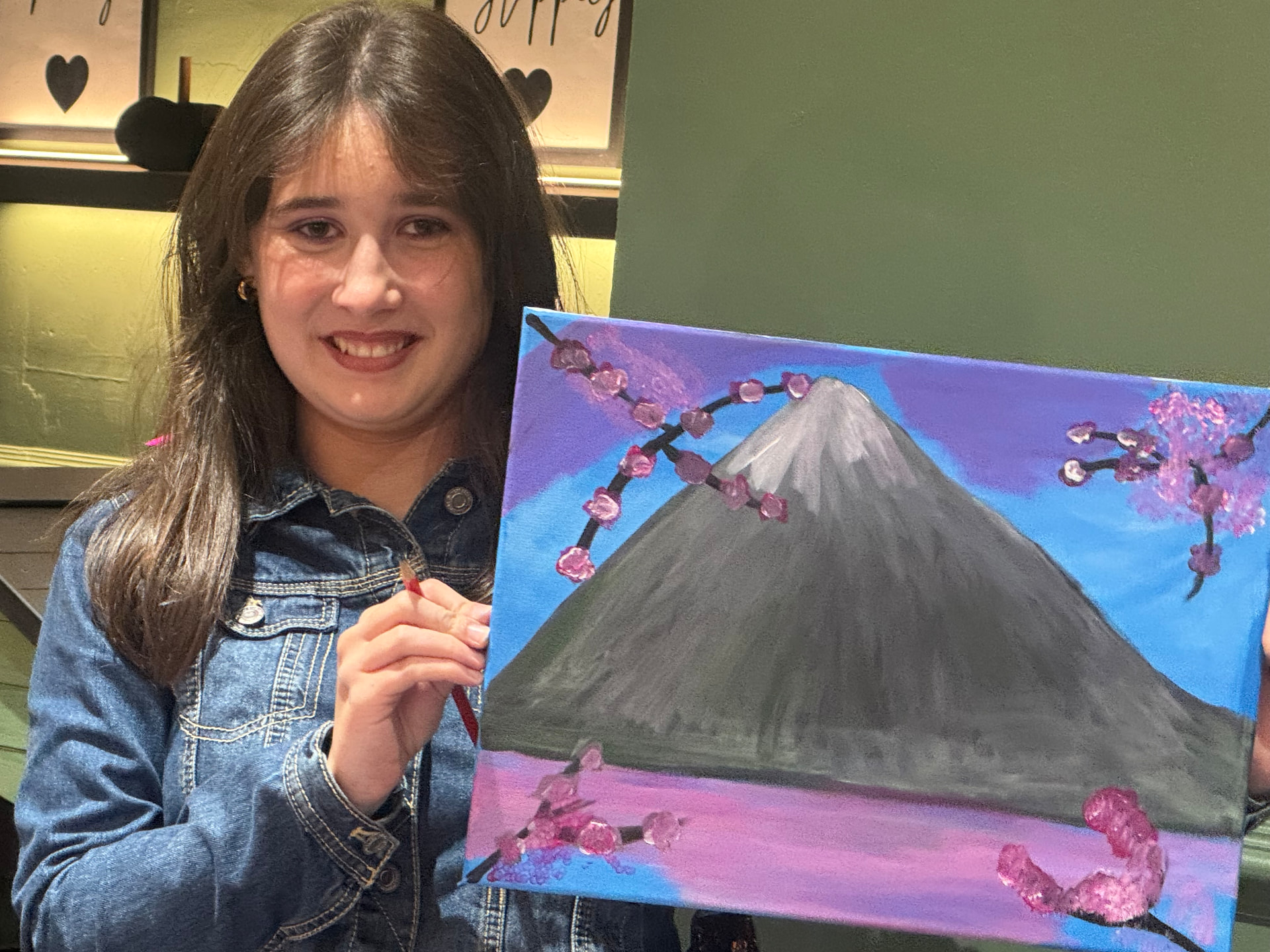 A young woman in a denim jacket holding a paintbrush beside her colourful Mount Fuji painting at the Newry social group.