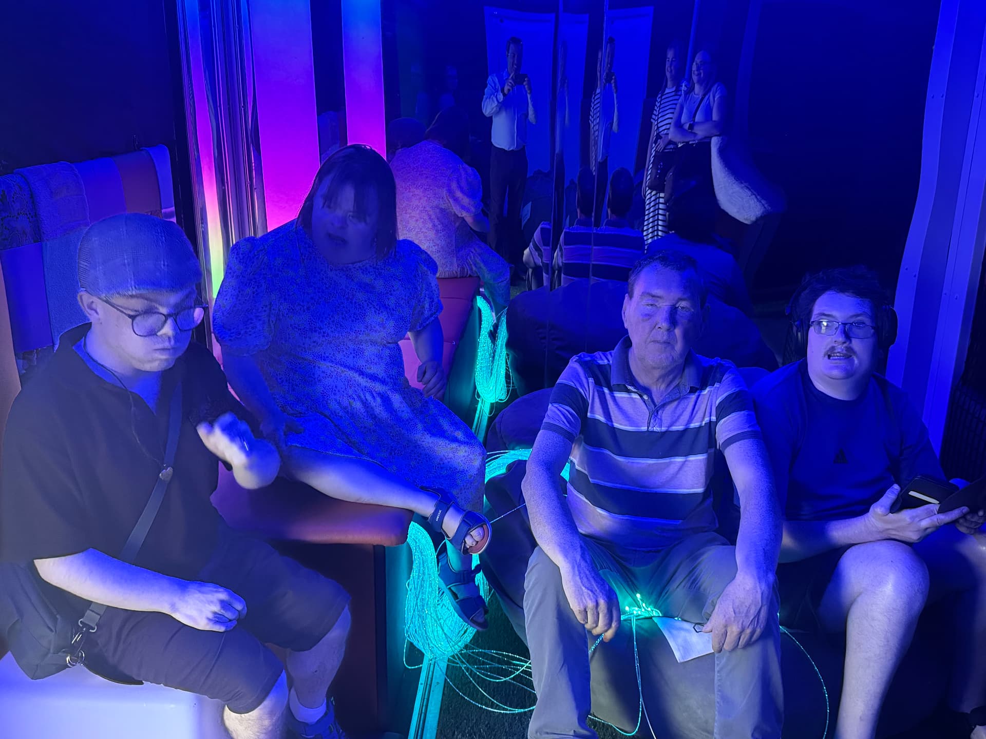 Young people and adults relaxing on soft seating in the softly lit mobile sensory room with fibre optic lights.