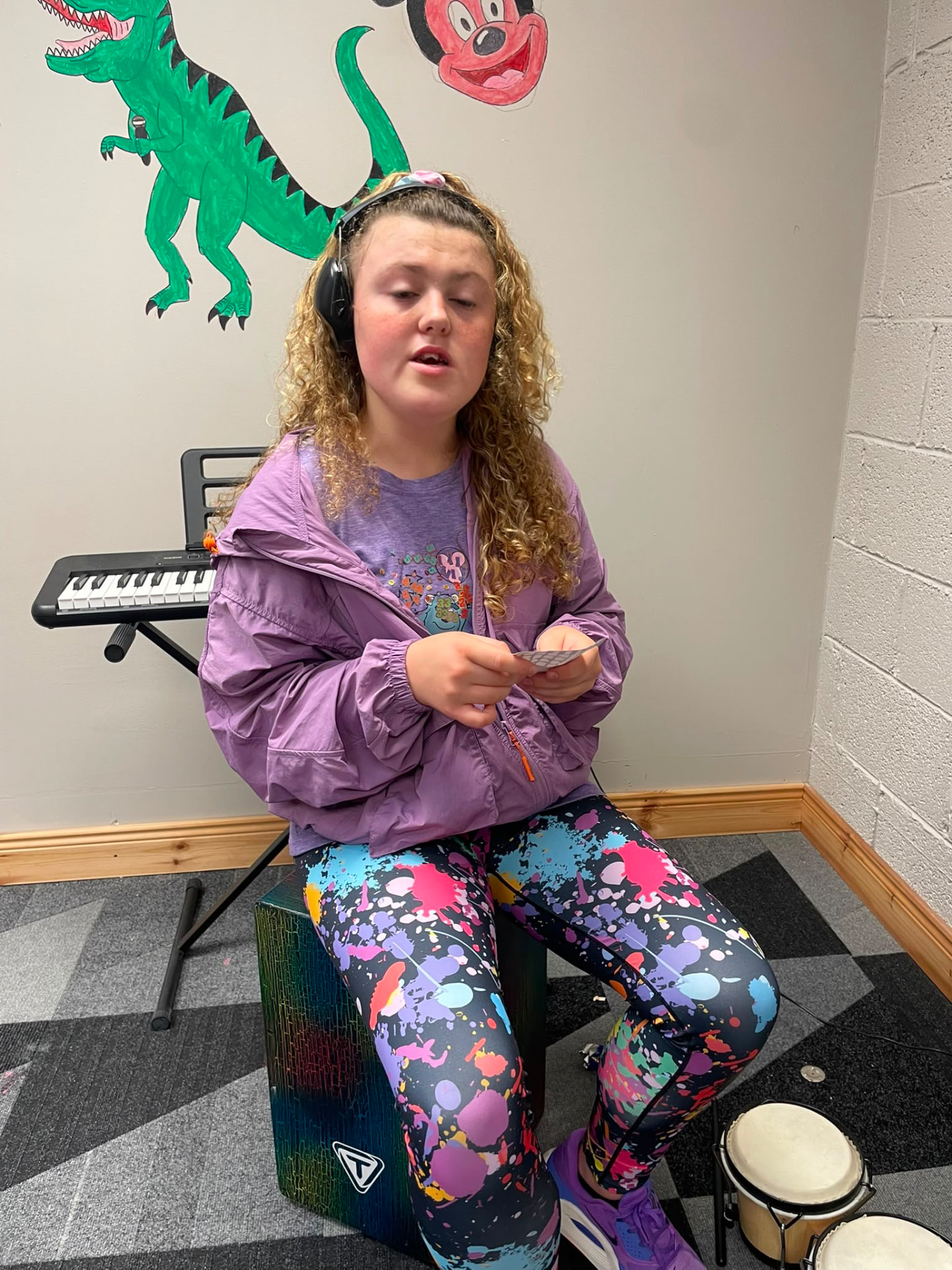 A girl wearing ear defenders playing a cajon drum at the ASD Ireland summer camp.
