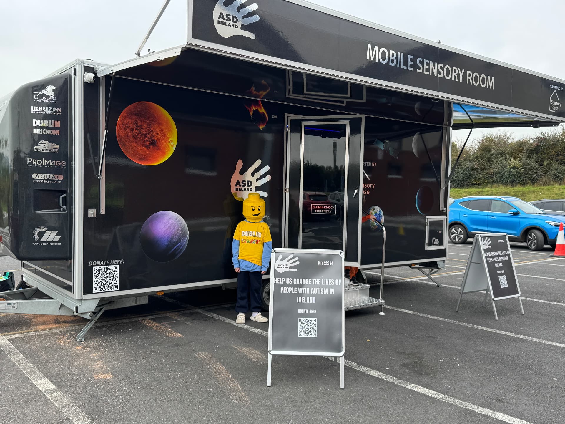 The ASD Ireland mobile sensory room with its awning open and supporter logos on the side, with a person in a yellow brick character costume at the door.